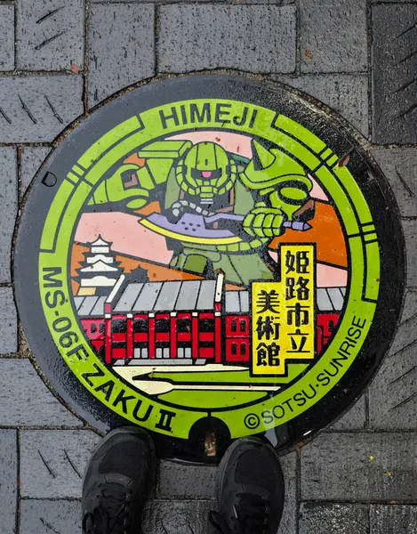 Kolorowa pokrywa studzienki w Himeji z wizerunkiem robota Zaku II na tle zamku i muzeum sztuki